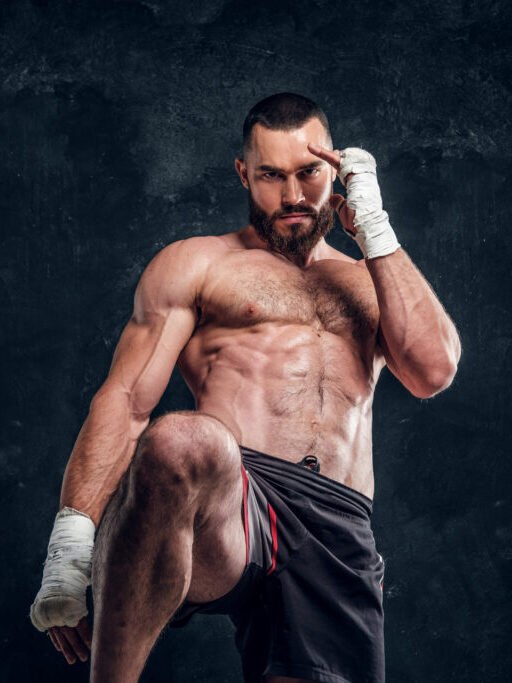 Strong muscular fighter is showing his punch while posing for photographer at dark photo studio.